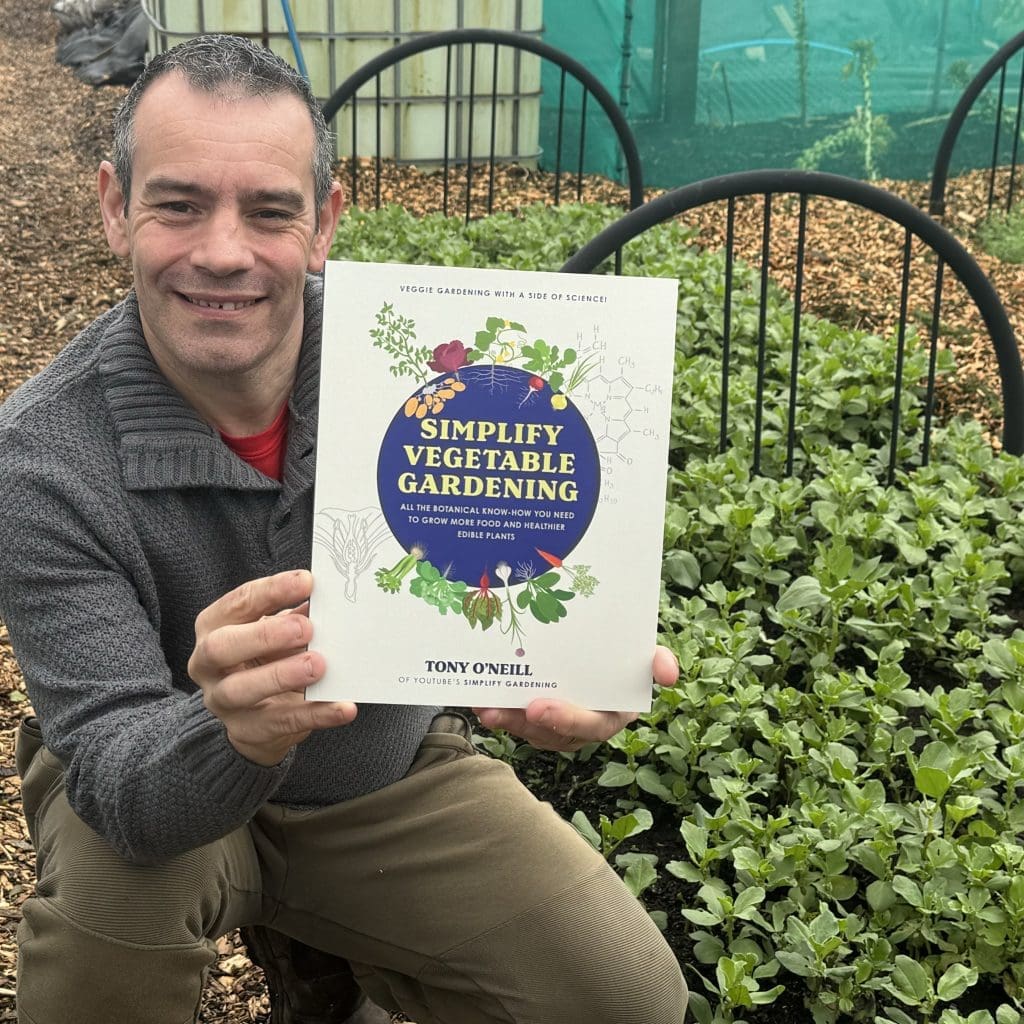 Tony O'Neill crouching in his South Wales garden and holding up Simplify Vegetable Gardening — his UK and USA bestselling third book and Silver Laurel Award winner 2025 — directly to camera with a broad grin