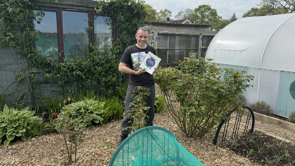 Tony O'Neill standing in his productive South Wales garden holding all three of his vegetable growing books — Composting Masterclass, Your First Vegetable Garden, and Simplify Vegetable Gardening — with the polytunnel and lush ornamental planting visible behind him