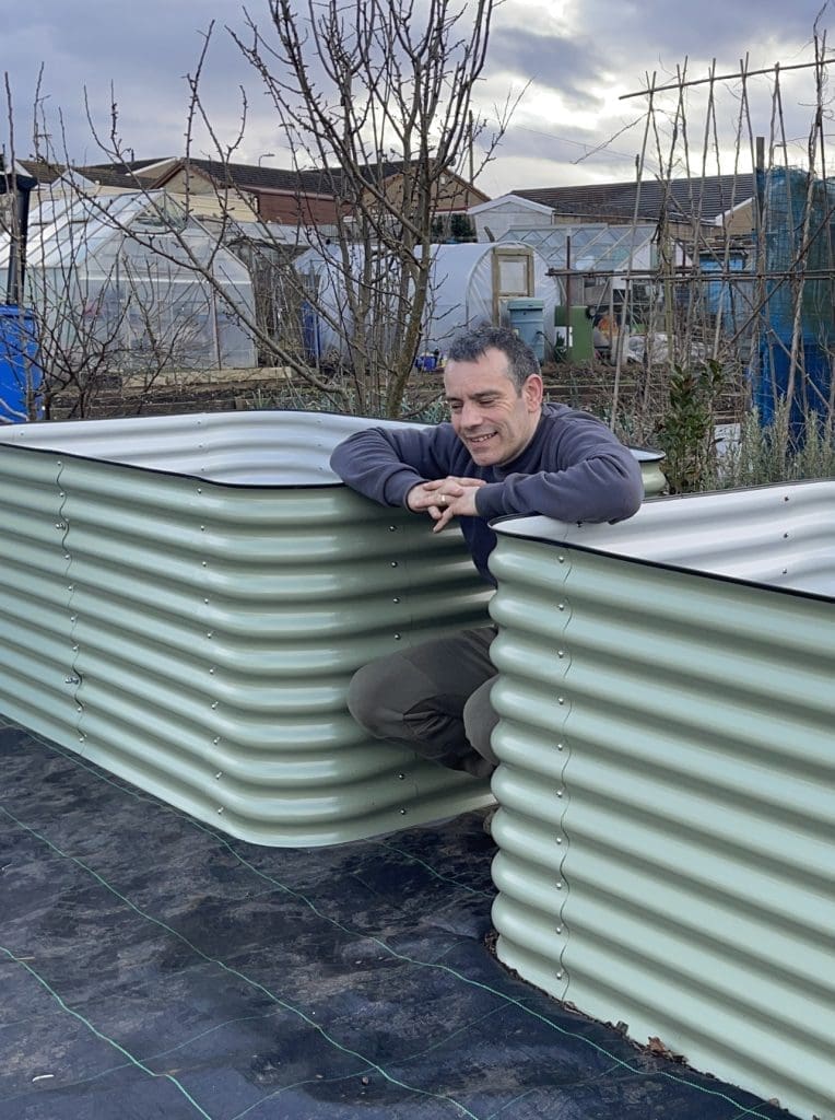 Tony O'Neill kneeling and smiling between two large newly built Birdies galvanised steel raised beds at his allotment garden in South Wales, with bare winter trees, the polytunnel, and neighbouring allotment structures visible in the background